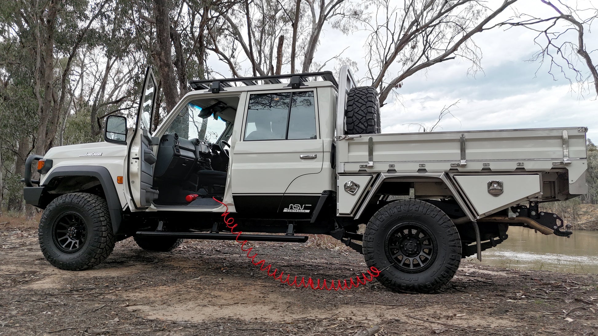 White off-road vehicle with a flatbed on a dirt road surrounded by trees.