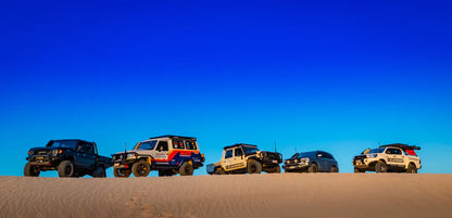 Five off-road vehicles parked on a sand dune with a clear blue sky.